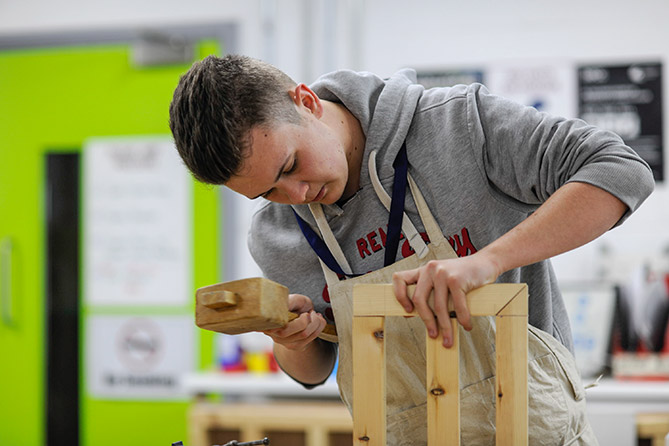 A young man in a joinery workshop uses a mallet to tap woodwork pieces into place 